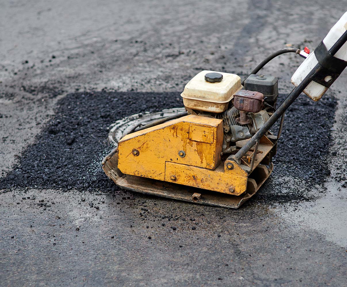 Close-up of a vibratory plate compactor being used to repair a pothole on an asphalt surface.